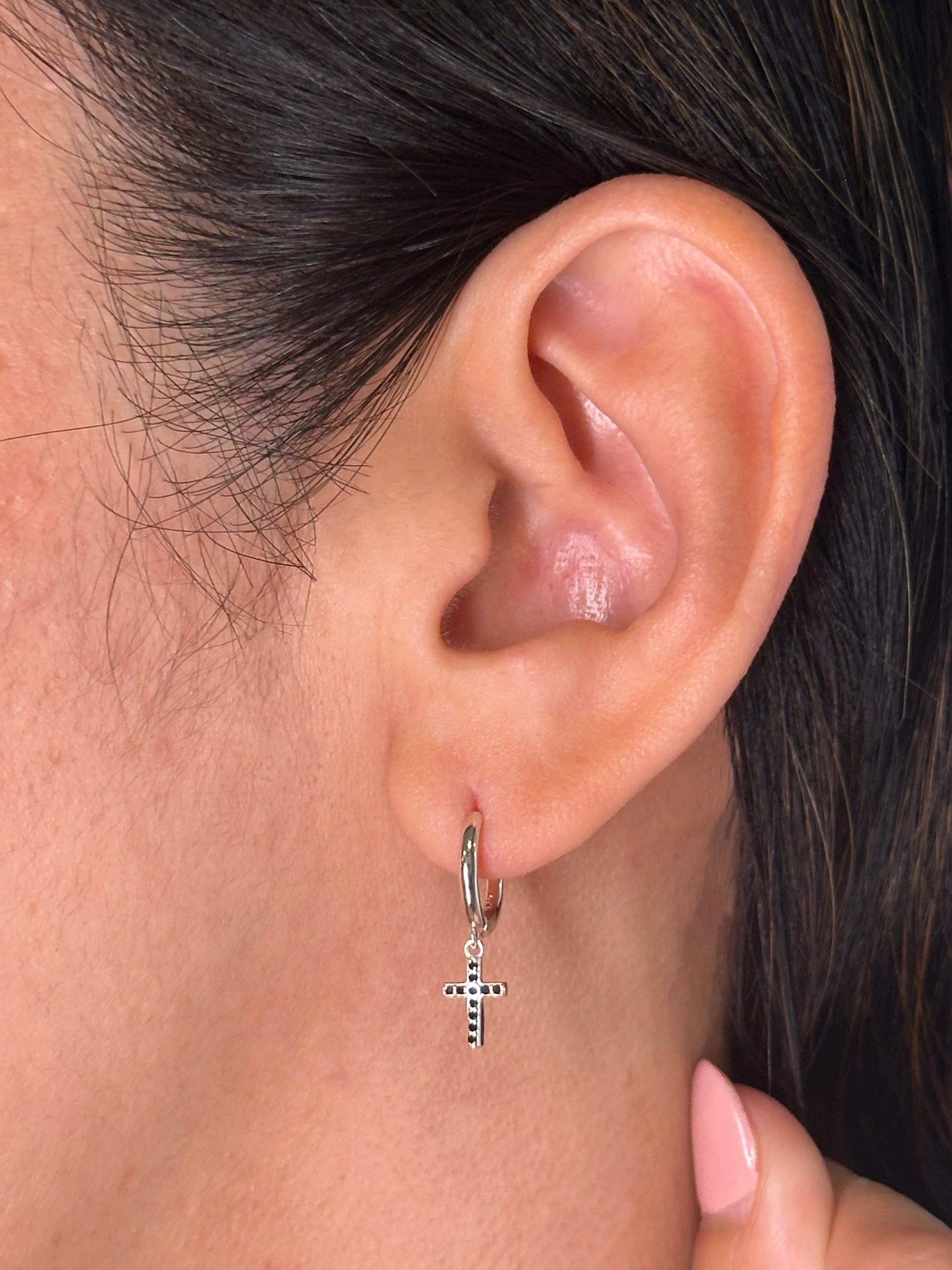 Close-up of a woman's ear wearing a sterling silver huggie earring with a cross charm, a timeless design perfect for casual or formal wear.