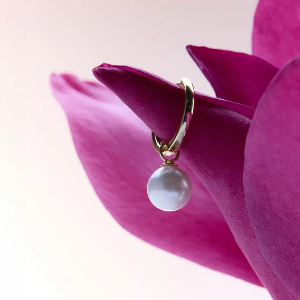 Detailed view of a yellow gold plated hinged hoop earring with a lustrous white pearl drop, elegantly set against a pink flower.