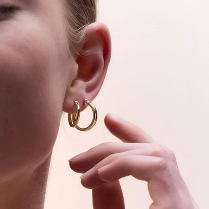 Yellow gold plated round tube hoop earrings worn, paired with a delicate hand gesture, emphasising their timeless elegance.