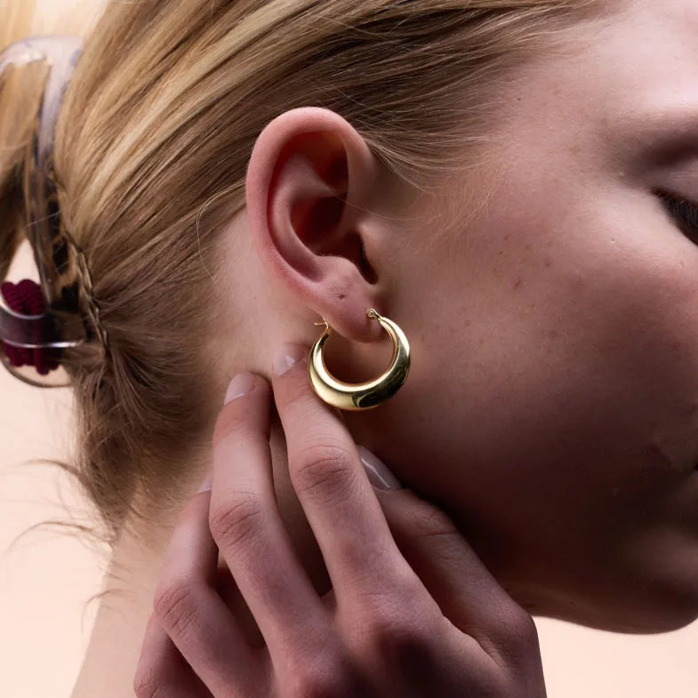 Model adjusting gold-plated tapered oval hoop earrings, emphasising their lightweight construction and refined design.