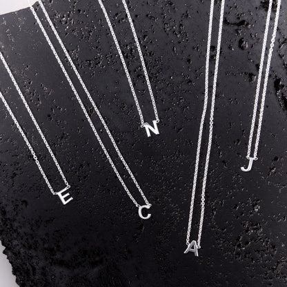 Multiple sterling silver initial necklaces ("C," "E," "N," "A," "J") displayed on a black textured surface, highlighting their timeless and versatile designs.