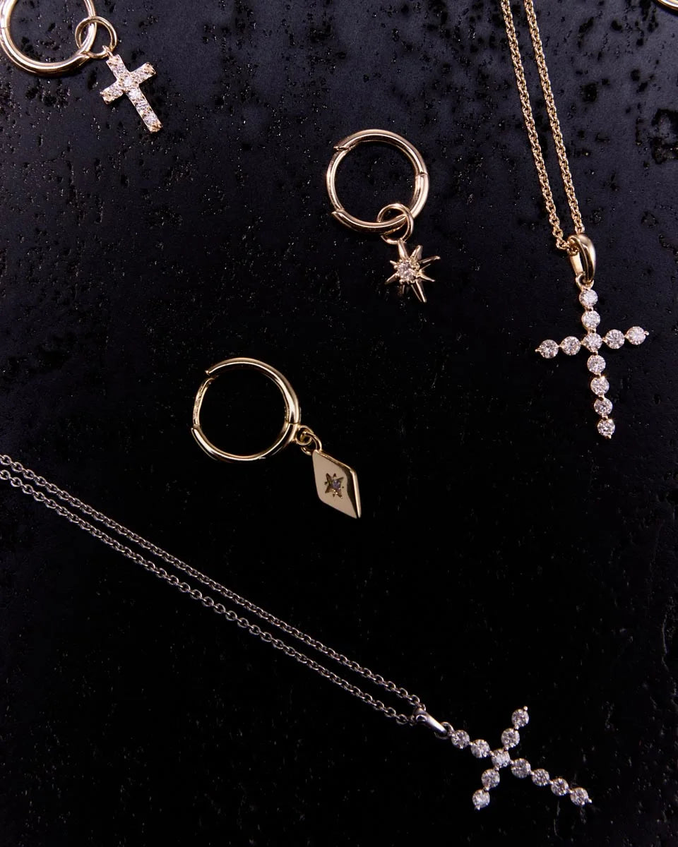 Flat-lay of 9k yellow gold jewellery featuring a diamond cross pendant on a chain, star and tag charms on hoop earrings, displayed on a textured black background.