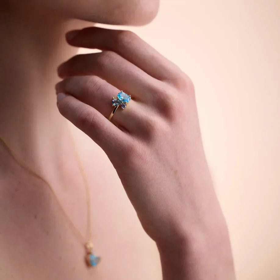 Close-up of a hand adorned with a gold-plated gemstone ring featuring London blue topaz and amethyst, paired with a matching necklace.