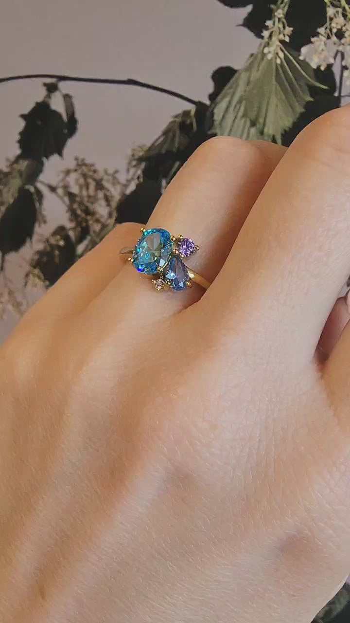 Close up of elegant gold-plated ring featuring a brilliant oval London blue topaz, a vibrant amethyst, and a sparkling cubic zirconia in a sophisticated cluster design worn on the ring finger of the left hand.